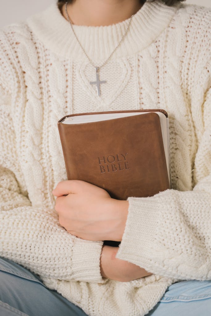 An individual in a knitted sweater holds a Holy Bible and wears a cross necklace, symbolizing faith.