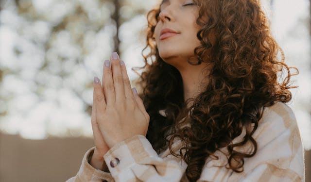 Serene woman with curly hair clasping hands in prayer outdoors.