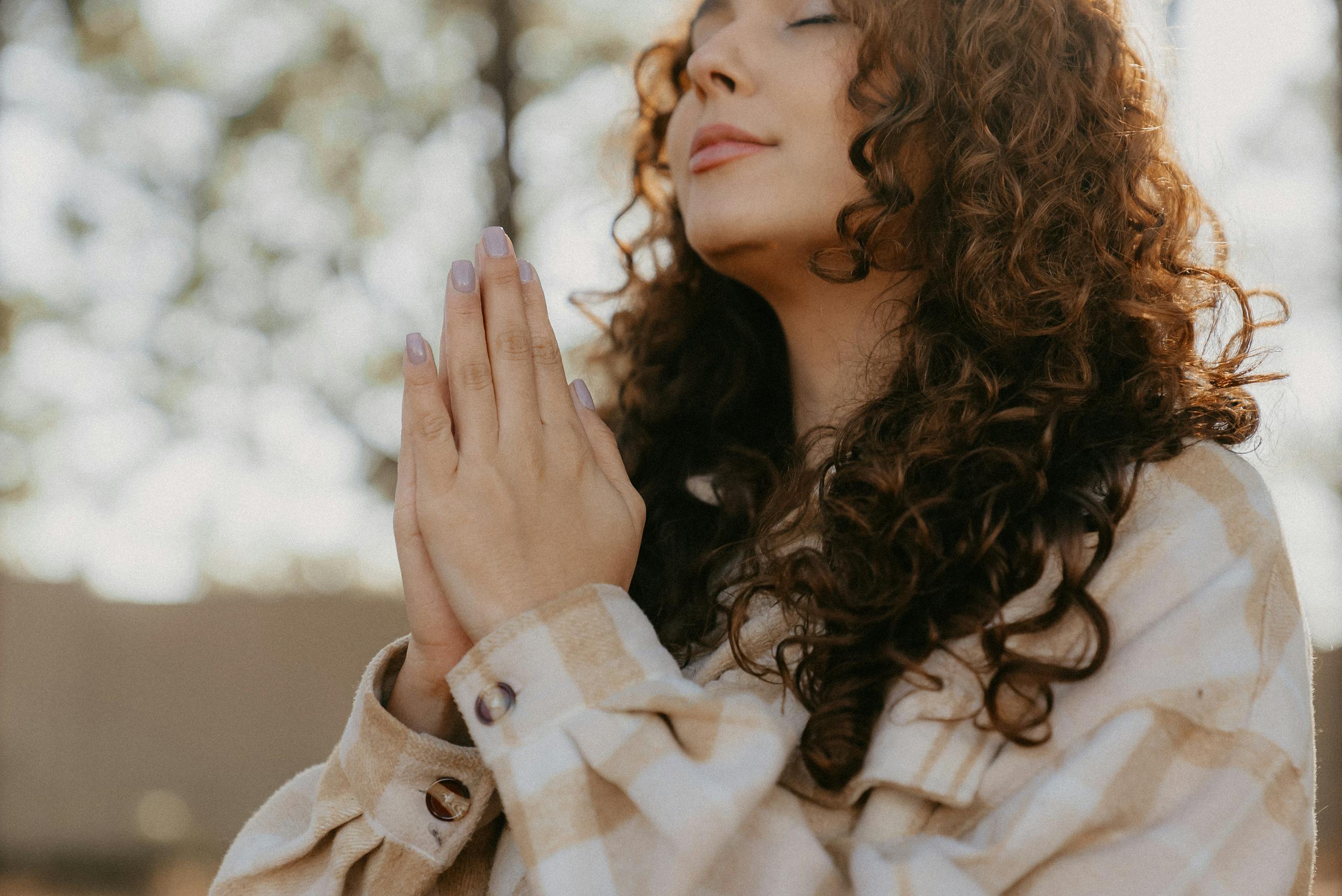 Serene woman with curly hair clasping hands in prayer outdoors.