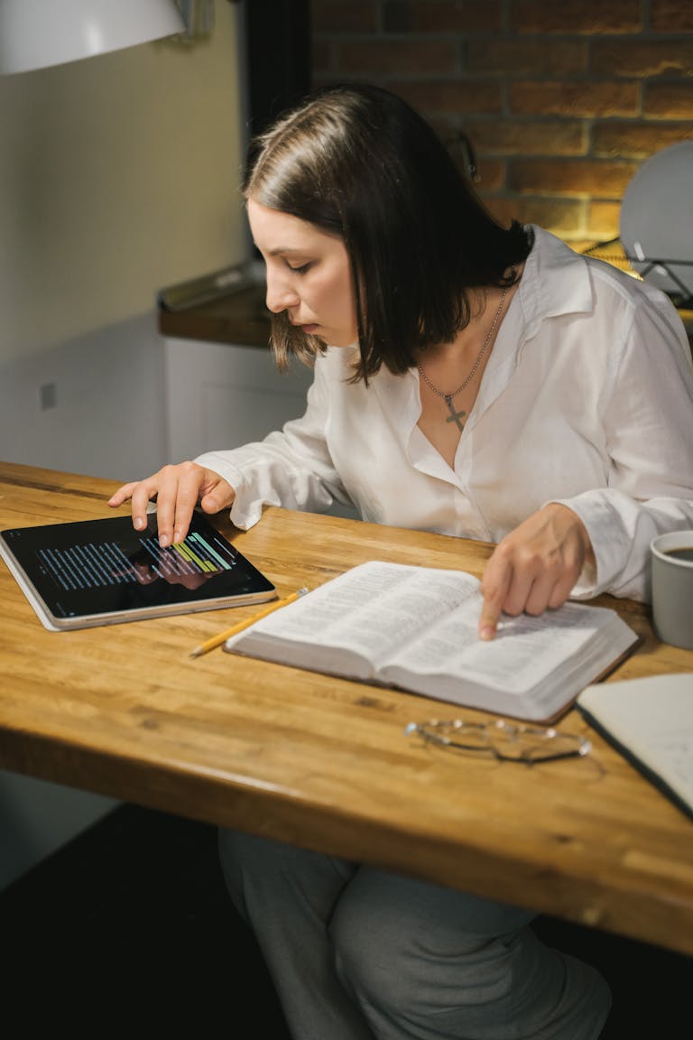 A woman studies the Bible at a wooden table, combining printed and digital texts.
