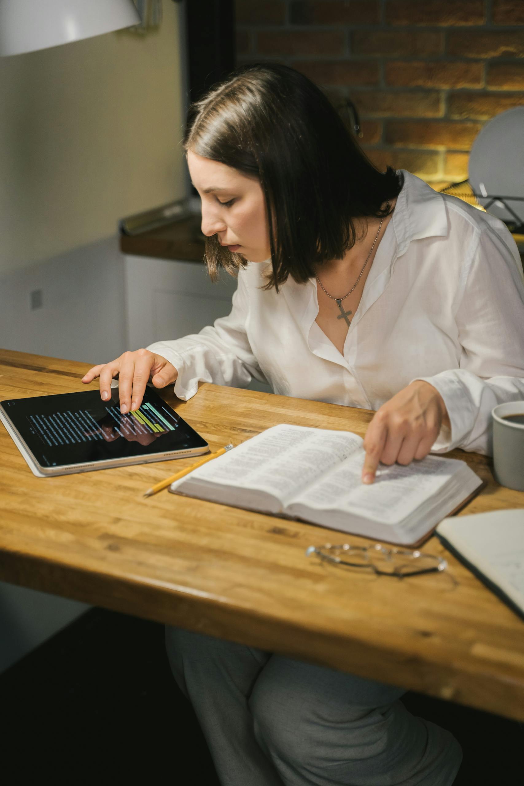A woman studies the Bible at a wooden table, combining printed and digital texts.