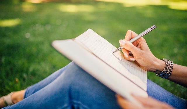 Person writes in a notebook on a sunny day. Focus on hands and pen outdoors.