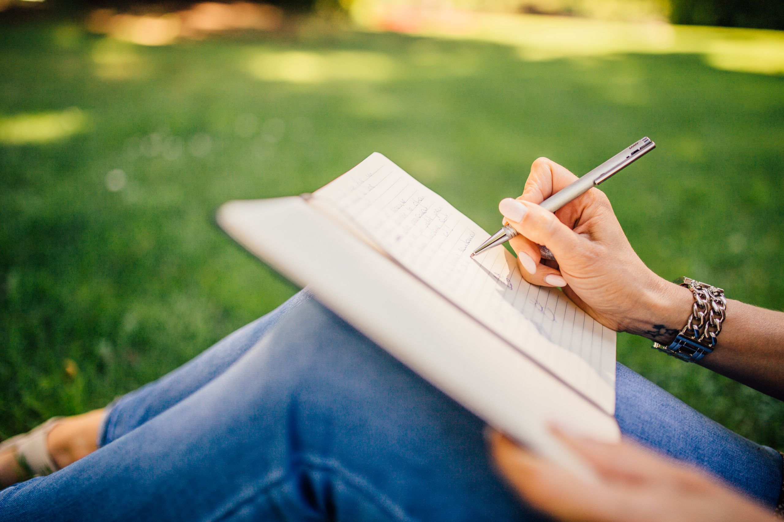 Person writes in a notebook on a sunny day. Focus on hands and pen outdoors.