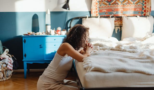 A woman kneeling by her bed in prayer, in a warmly lit, cozy bedroom setting.