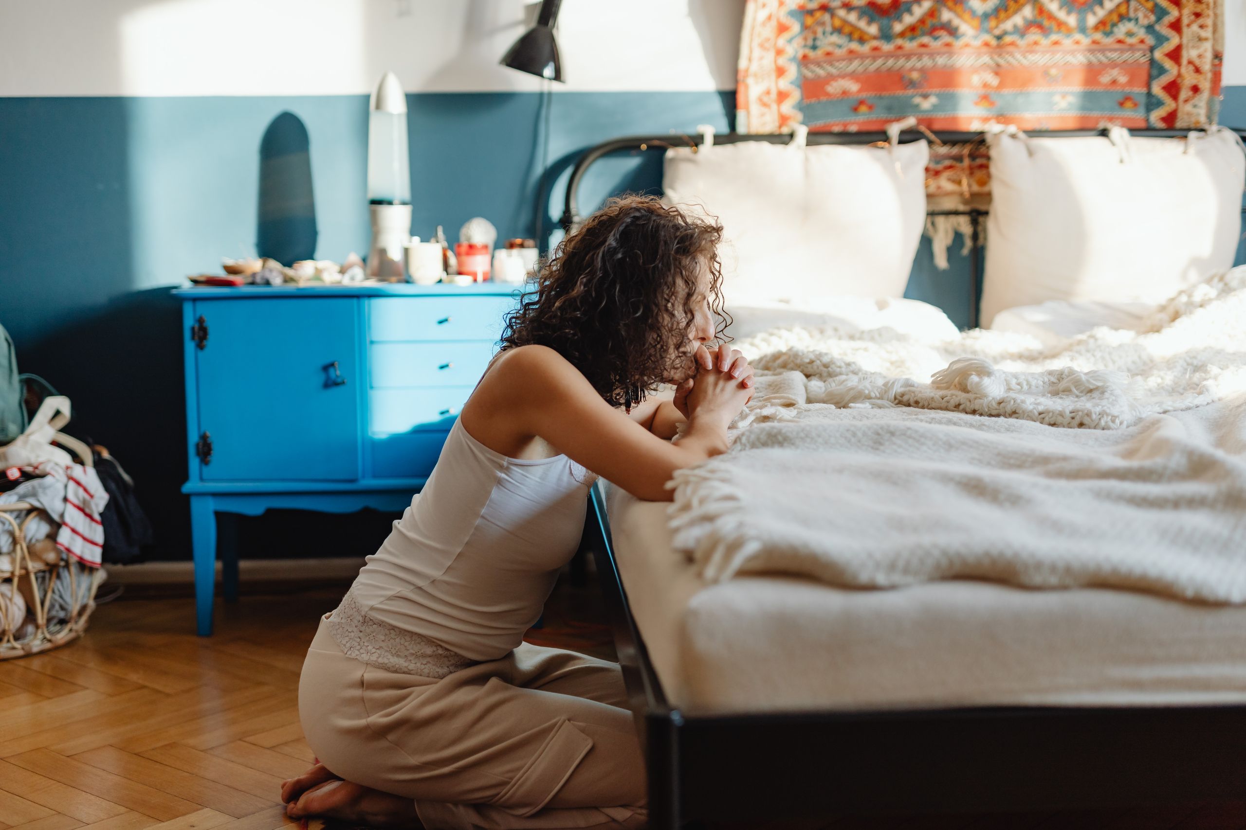 A woman kneeling by her bed in prayer, in a warmly lit, cozy bedroom setting.